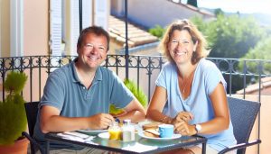 Un couple souriant, assis à une table en plein air sur une terrasse ensoleillée, en train de prendre leur petit déjeuner.